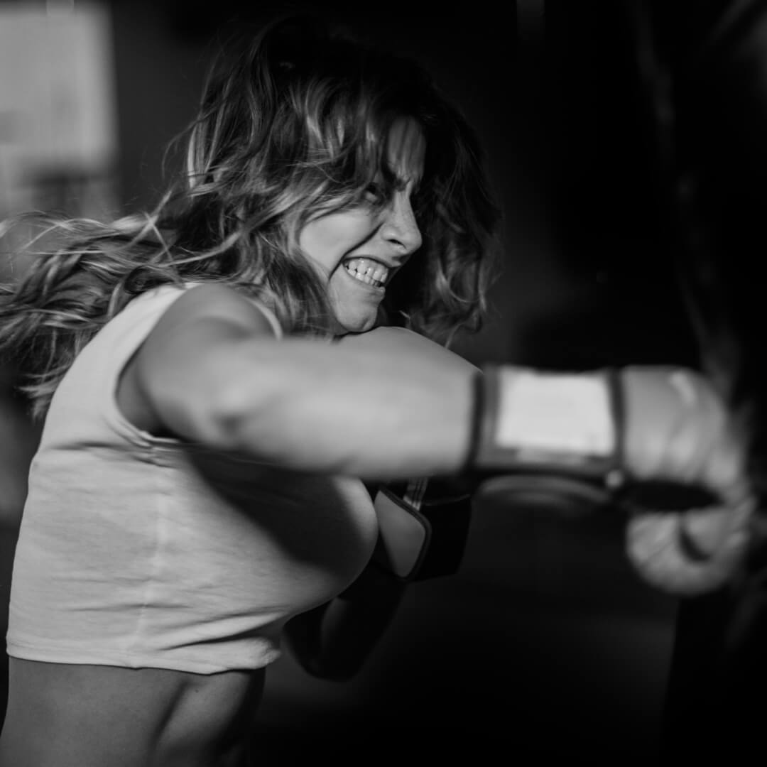 A black and white lifestyle photograph of a woman punching a heavy bag in a gym, wearing a white tank top and boxing gloves. She is an older athlete that stays healthy with Battle Balm all natural & organic topical pain cream