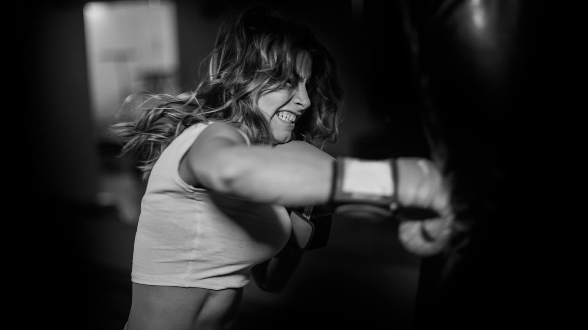 A black and white lifestyle photograph of a woman punching a heavy bag in a gym, wearing a white tank top and boxing gloves. She is an older athlete that stays healthy with Battle Balm all natural & organic topical pain cream