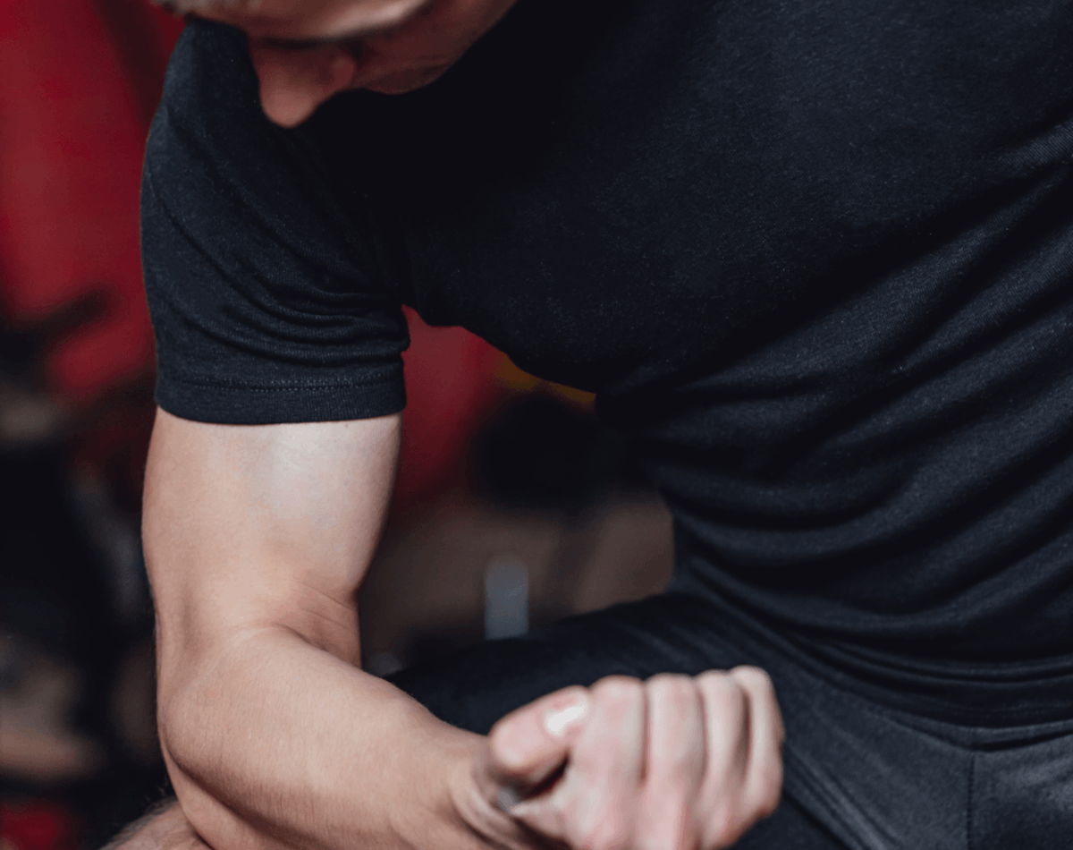 Athlete assessing elbow pain at gym and preparing to apply Battle Balm topical pain relief to musculature surrounding elbow zoomed in 10%